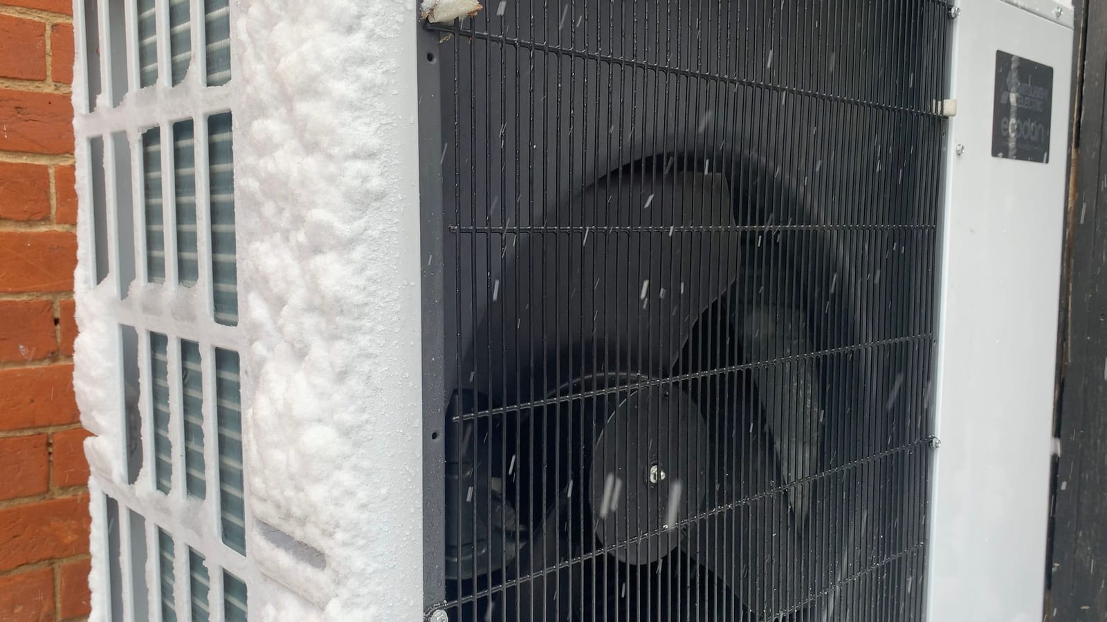 The outdoor unit of an air-source heat pump beside a brick wall, with snow built up on its side grille while snow falls.